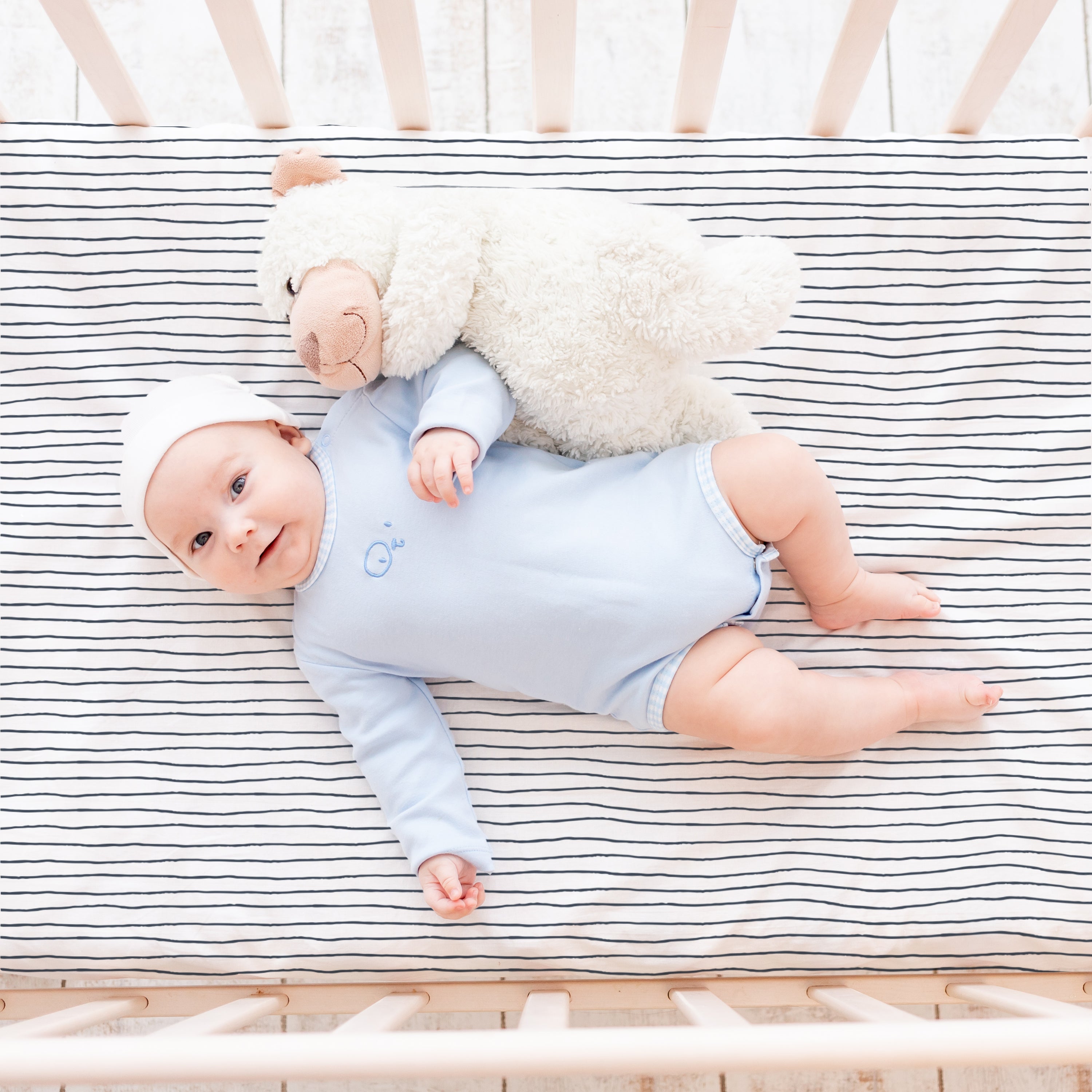 A baby in a blue onesie lies in a Makemake Organics crib with a Cobi Blue Stripes fitted sheet and pillowcase, accompanied by a plush teddy bear on its side. The baby is looking up and smiling, wearing a white headband.