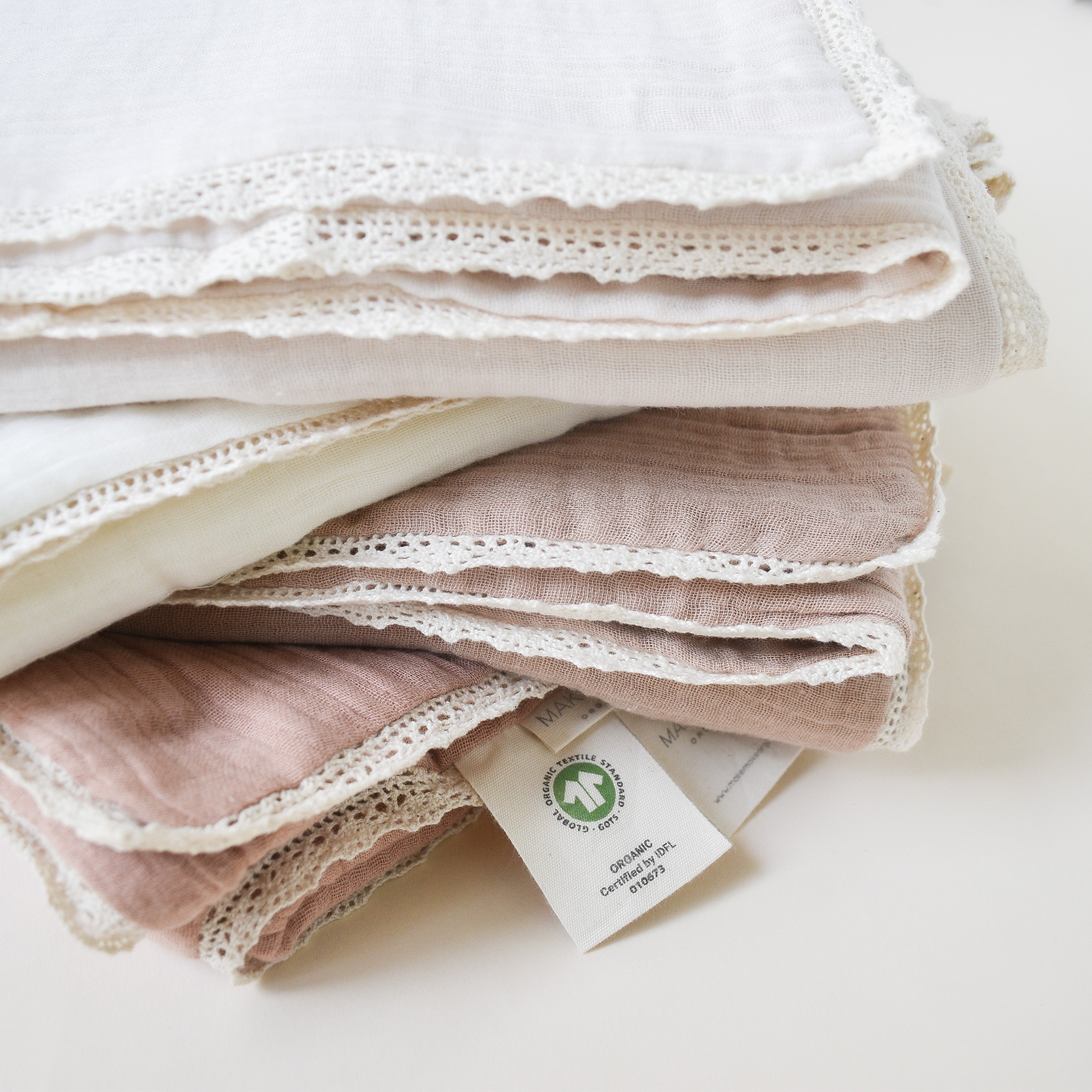 A stack of neatly folded Makemake Organics Organic Cotton Muslin Blanket - Blush Oat + Ivory Lace in shades of white and pink, each edged with delicate lace, displaying a green certification tag.