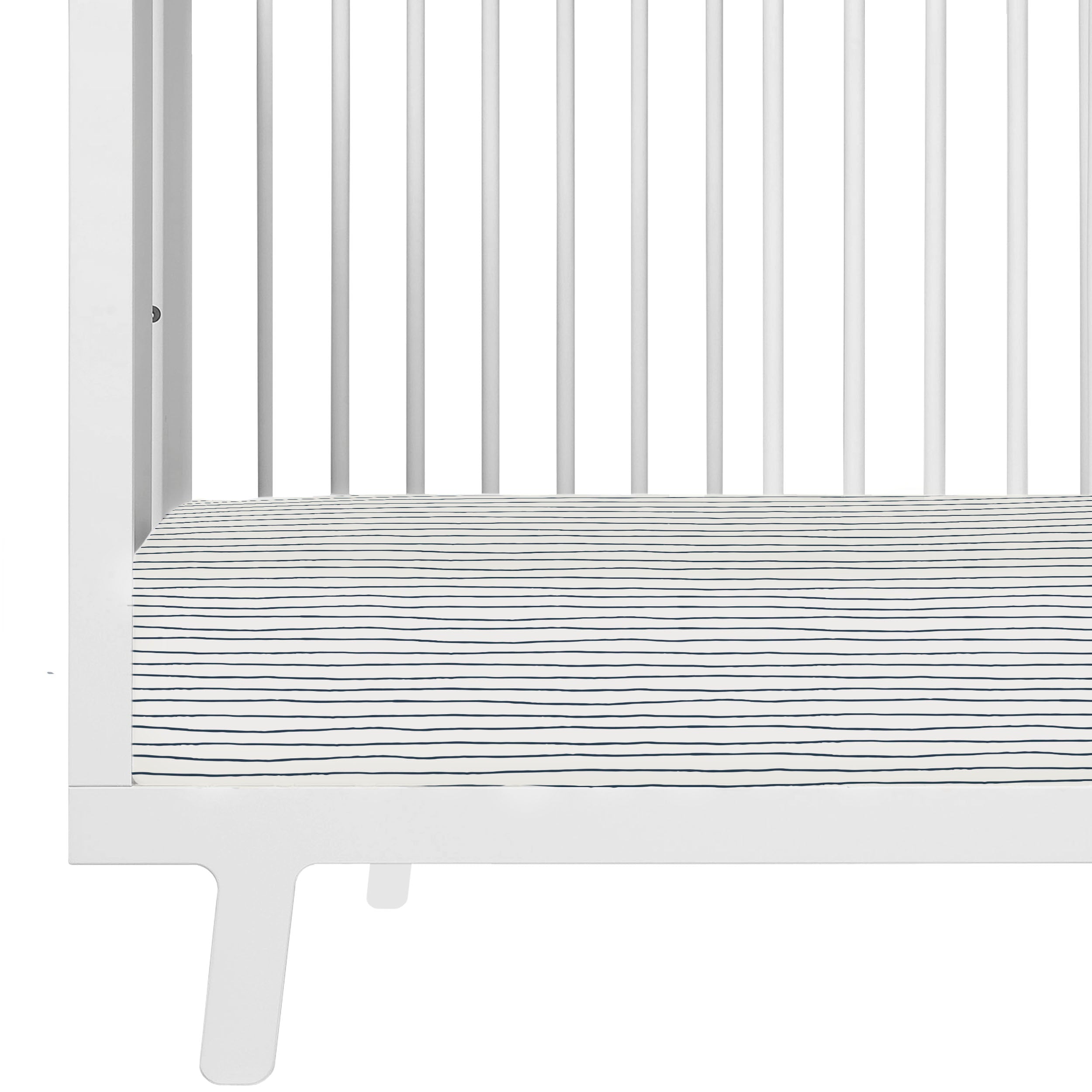 Close-up of a white Makemake Organics baby crib with a Cobi Blue Stripes fitted sheet and pillowcase visible through the vertical slats. The crib showcases a clean, modern design with a single visible leg.