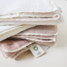 A stack of neatly folded Makemake Organics Organic Cotton Muslin Blankets with delicate lace trim in shades of white and soft pink on a light background.