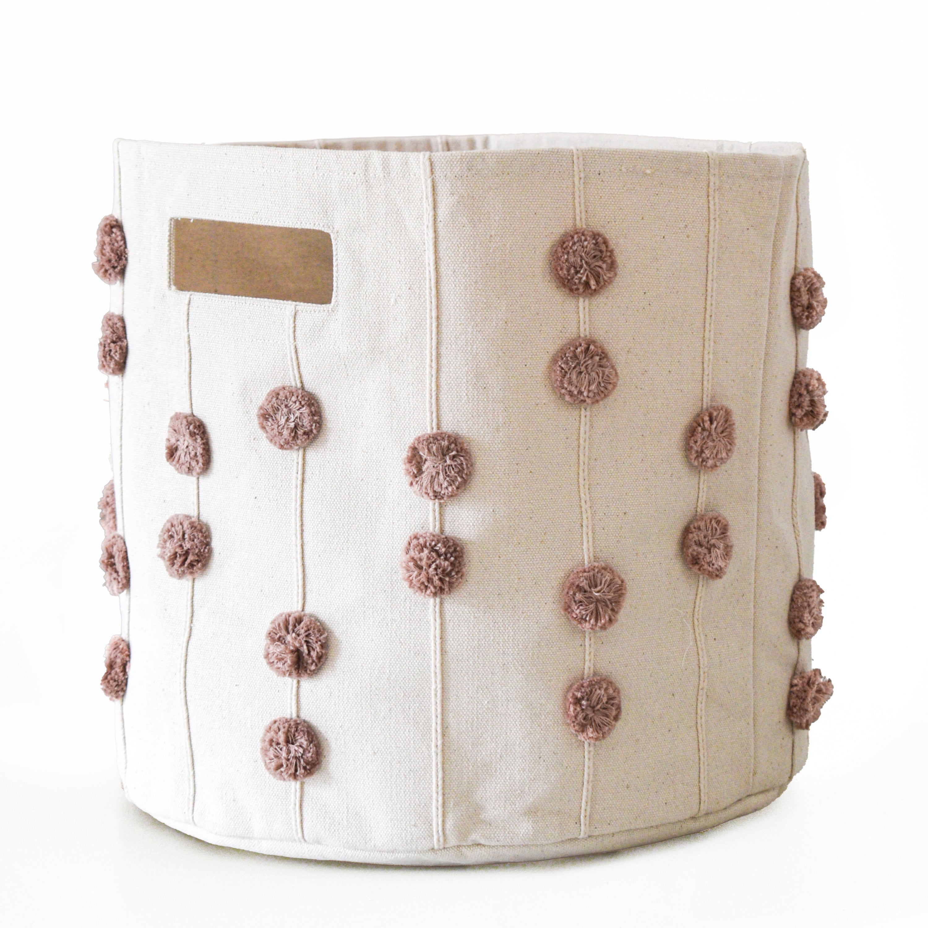 A beige fabric Storage Basket Pompom Pecan decorated with rows of small, soft, pink pom-poms and a small rectangular label at the top. the background is plain white. Brand Name: Makemake Organics