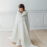 A young girl wrapped in an oversized Organic Cotton Muslin Blanket - Ivory + Ivory Lace from Makemake Organics, standing thoughtfully with her head down in a bright room with white paneled walls.
