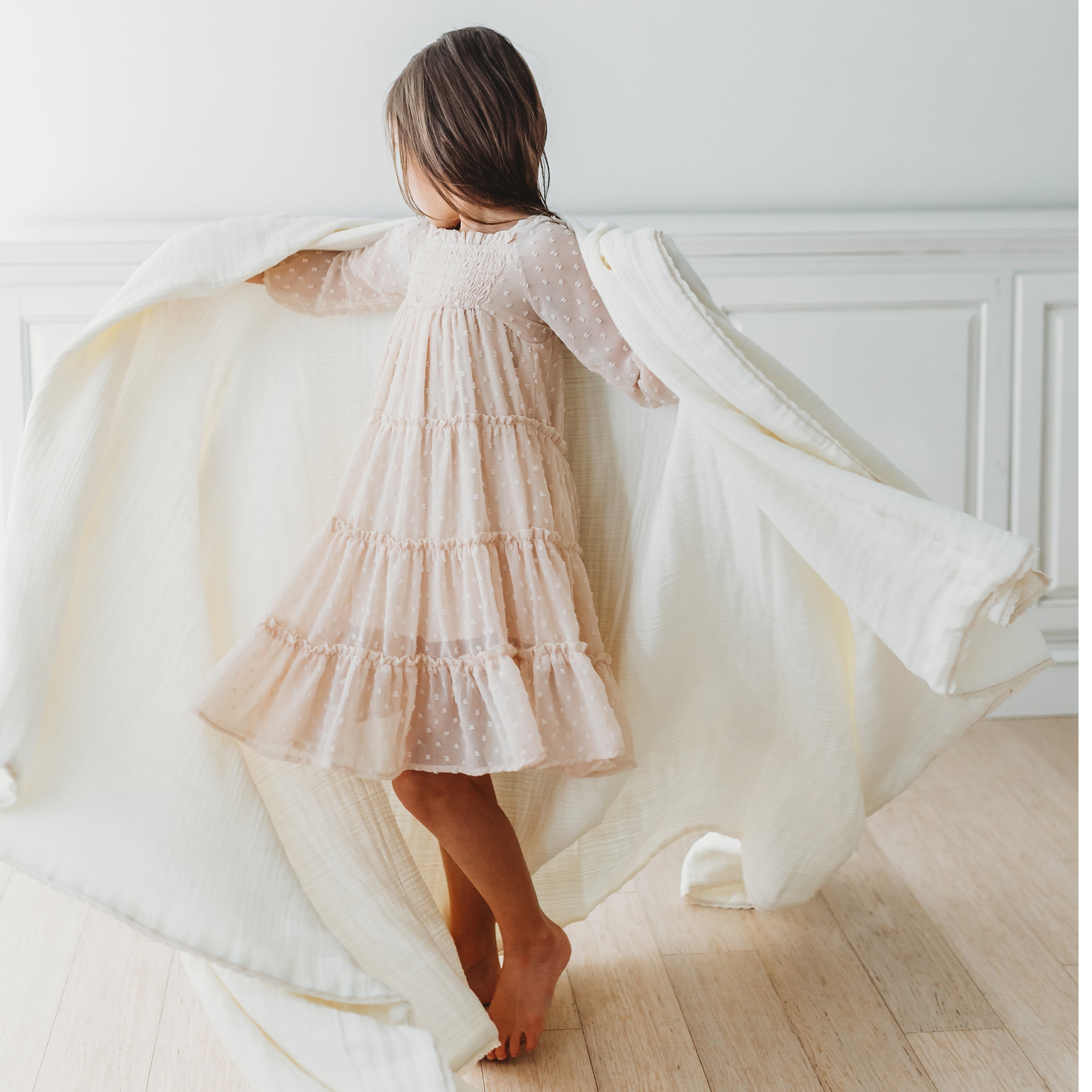 A young girl in a flowing beige dress twirls away from the camera in a bright room, her dress and Makemake Organics Organic Cotton Muslin Blanket - Ivory + Ivory Lace caught gracefully in mid-motion.