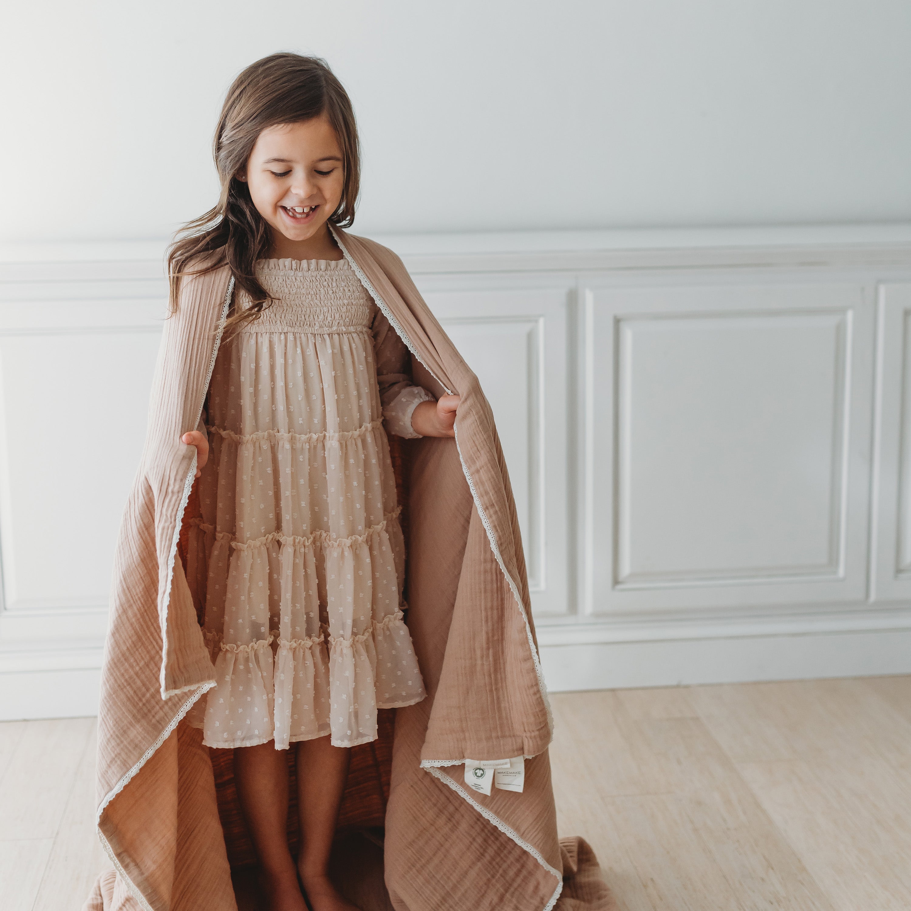 A young girl stands smiling, holding open a long, flowing Organic Cotton Muslin Blanket in Taupe + Natural Lace over a layered beige dress, in a bright room with white walls by Makemake Organics.