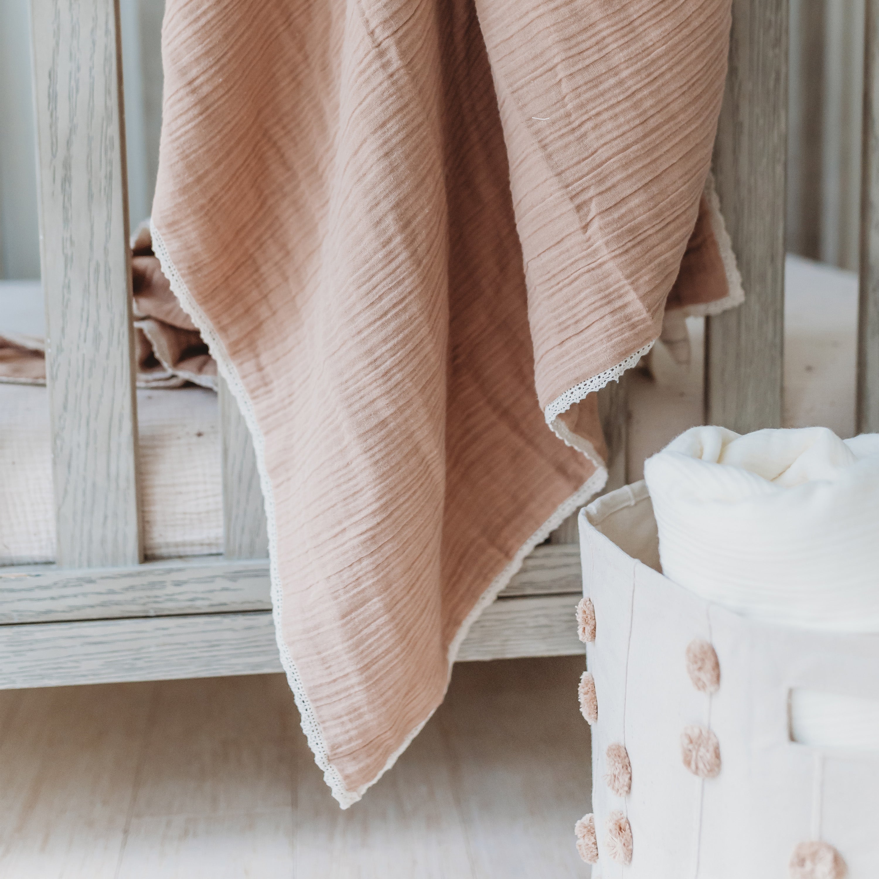 A soft pink Organic Cotton Muslin Blanket - Pecan + Natural Lace draped elegantly over a wooden ladder beside a white wicker basket with a fluffy white blanket, suggesting a cozy, serene nursery setting from Makemake Organics.