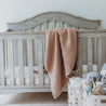 A cozy wooden baby crib with soft toys and Makemake Organics Organic Cotton Muslin Blanket - Taupe + Natural Lace in a nursery room, conveying a calm and comforting atmosphere.