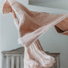 A woman in a flowing, textured pink dress captured mid-spin, with only her arm visible, tossing the skirt. the background features a soft-focus on a Makemake Organics Organic Cotton Muslin Blanket - Pecan + Natural Lace.