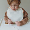 A toddler with a Makemake Organics Organic Muslin Bibs - Ivory & Taupe sitting in a highchair, examining small objects on the tray in front of him.