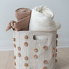 A Storage Basket Pompom Taupe from Makemake Organics containing neatly rolled cozy blankets in shades of cream and dusty pink, placed on a wooden floor against a plain wall.