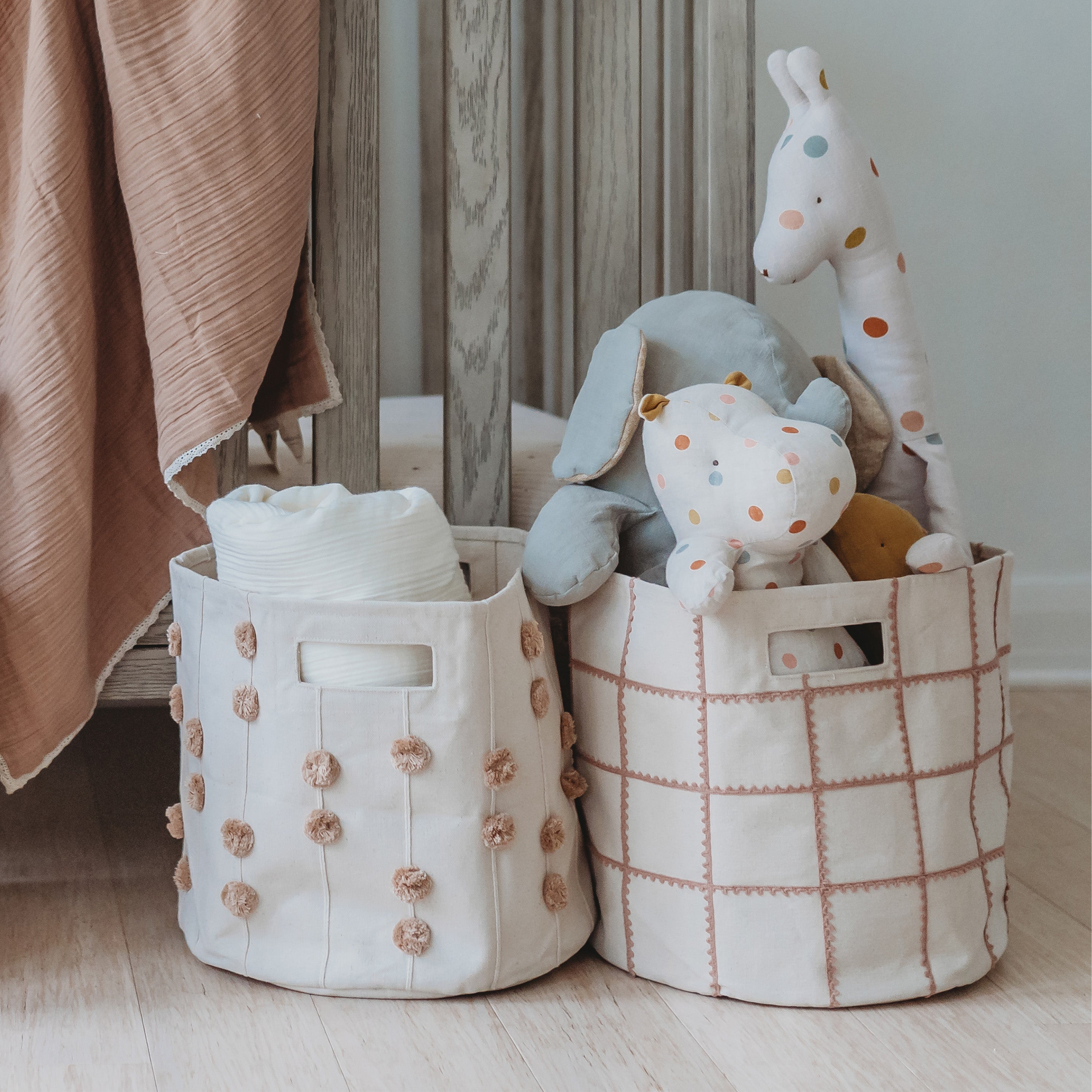 Two stylish Makemake Organics Storage Basket Mesh Lace - Pecan, filled with children's toys, including stuffed animals and soft blocks, situated next to draped pink fabric and a wooden backdrop.