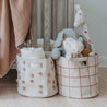 Two stylish Makemake Organics fabric Storage Basket Pompom Taupe filled with plush toys and baby blankets next to a wooden crib with a draped peach blanket.