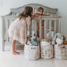 A young girl in a pink dress kisses a toddler boy on the cheek as he sits inside a wooden crib surrounded by Storage Basket Pompom Pecan from Makemake Organics.