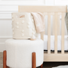 A stylish nursery room featuring a cream-colored padded ottoman with a wooden base, on which rests a beige baby backpack and a knit baby blanket with pompoms. A Makemake Organics Storage Basket Pompom Ivory is also visible in the room.