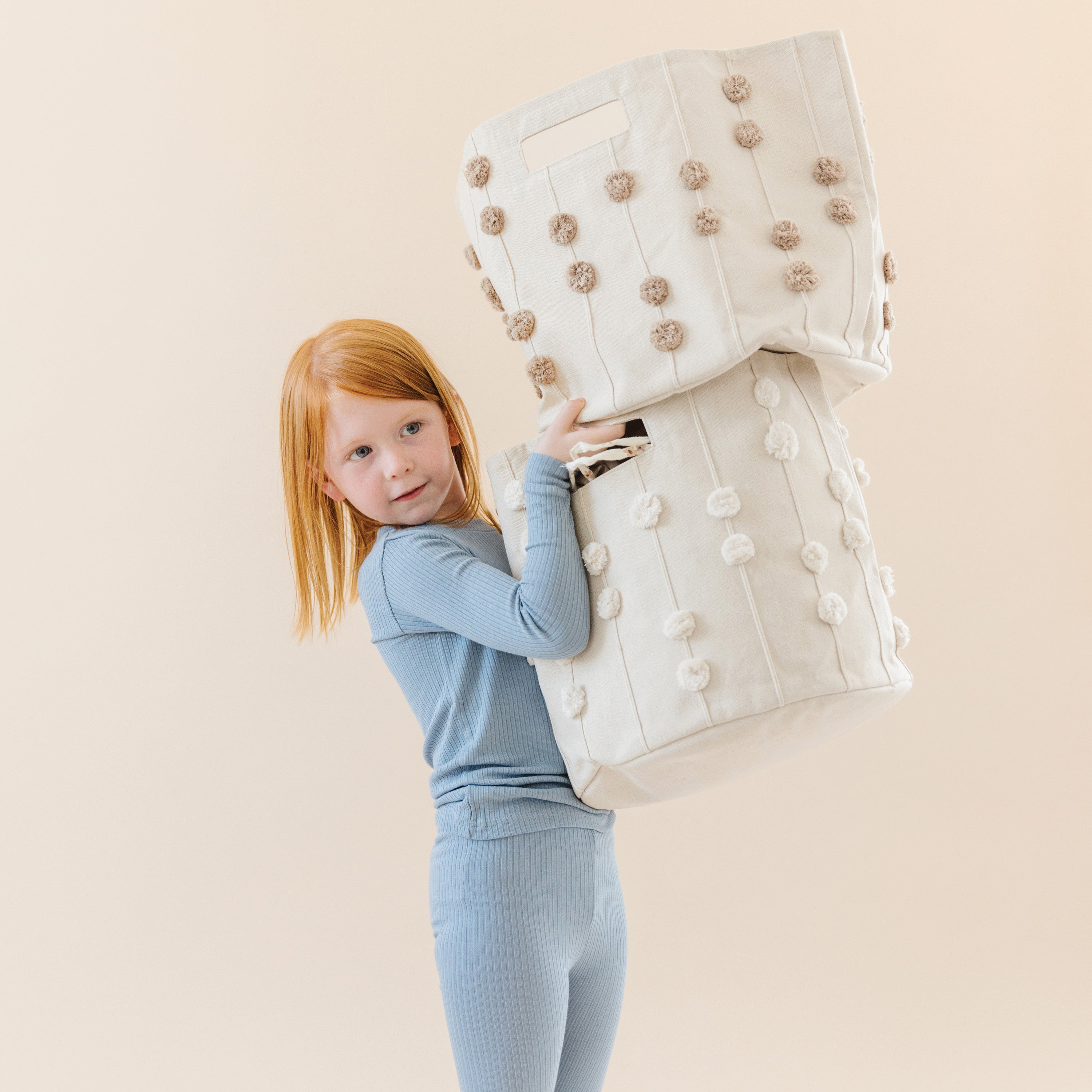A young girl with red hair, wearing a blue outfit, playfully holds up a large white Storage Basket Pompom Taupe from Makemake Organics against a light beige background.