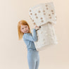 A young girl with red hair, wearing a blue outfit, playfully holds up a large white Storage Basket Pompom Taupe from Makemake Organics against a light beige background.
