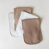 Three Organic Cotton Muslin Burp Cloths in ivory and taupe by Makemake Organics arranged overlapping on a light beige background. Each burp cloth has a visible label tag on the side.