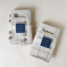 Two neatly folded beige Makemake Organics Storage Basket Mesh Lace - Taupe with labels displayed on a white background. Each caddy is adorned with decorative pom-poms along their edges.