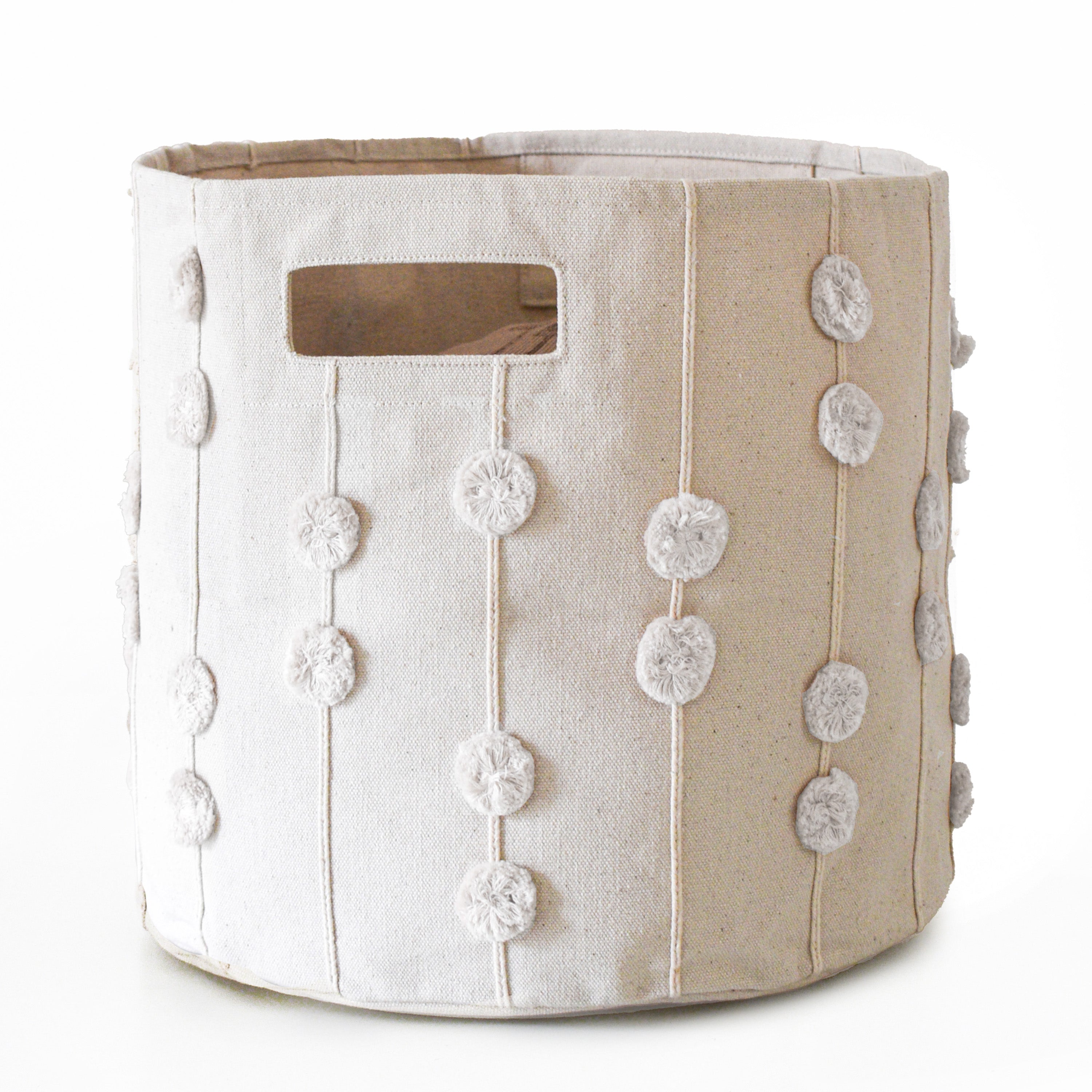 A Storage Basket Pompom Ivory by Makemake Organics with a window slot and vertical rows of decorative white pompoms against a white background.
