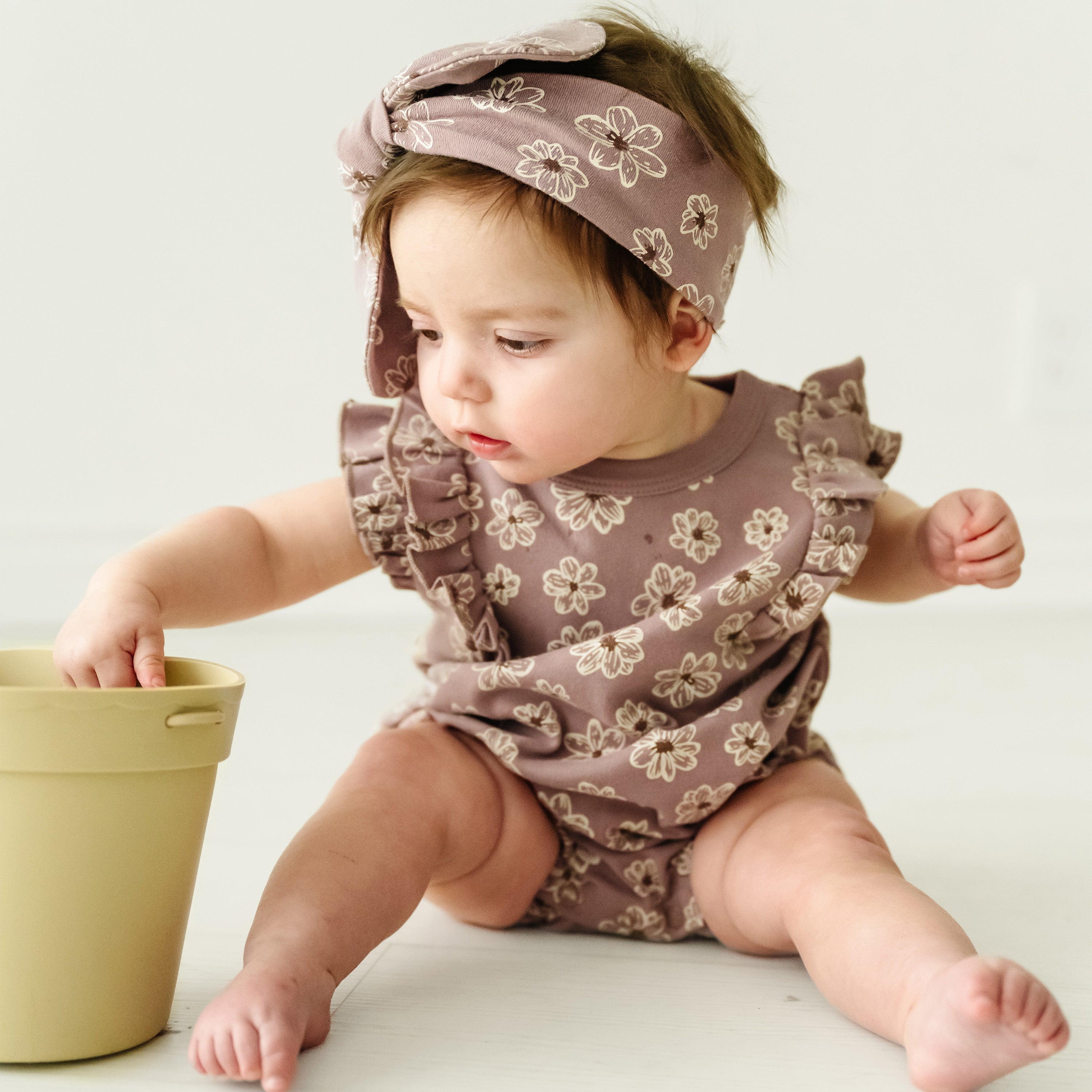 A toddler in a brown floral outfit and headband examines an Organic Flutter Bubble Onesie - Daisies container, sitting on a white surface with a curious expression.
