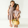 Two young toddlers in matching Makemake Organics Organic Flutter Dresses in Daisies stand holding a wicker basket with flowers, smiling in a brightly lit room.