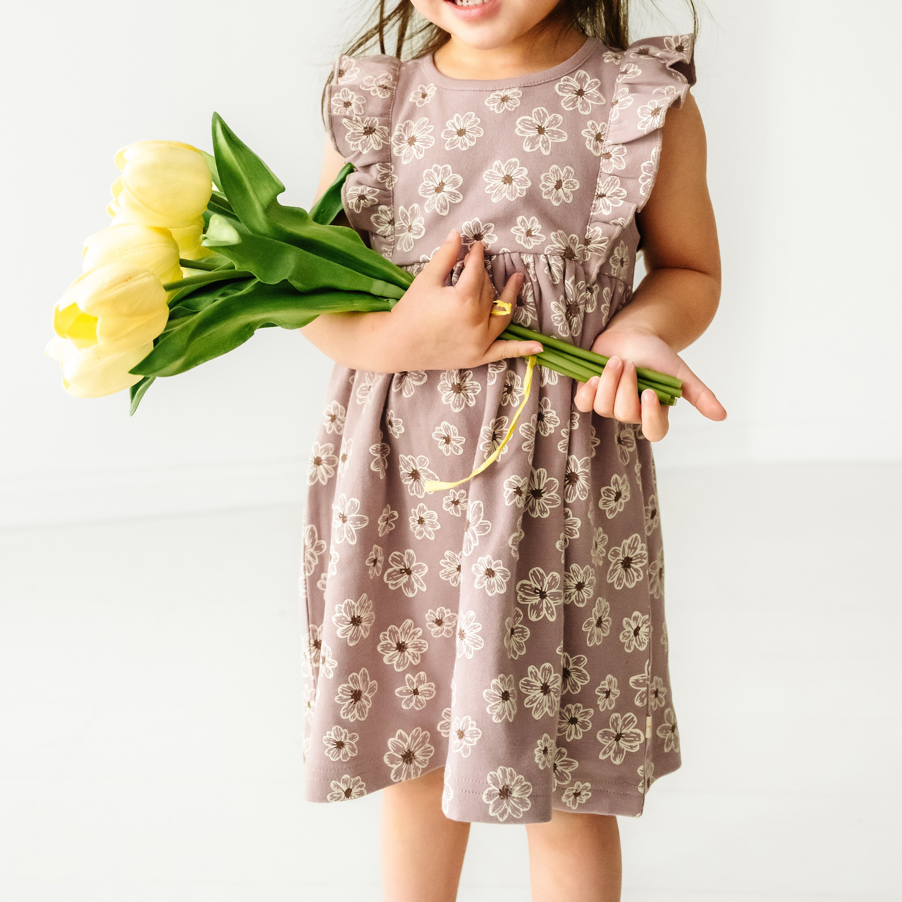 A young toddler in an Organic Flutter Dress - Daisies by Makemake Organics, holding a bouquet of yellow tulips, smiling while posing against a white background.