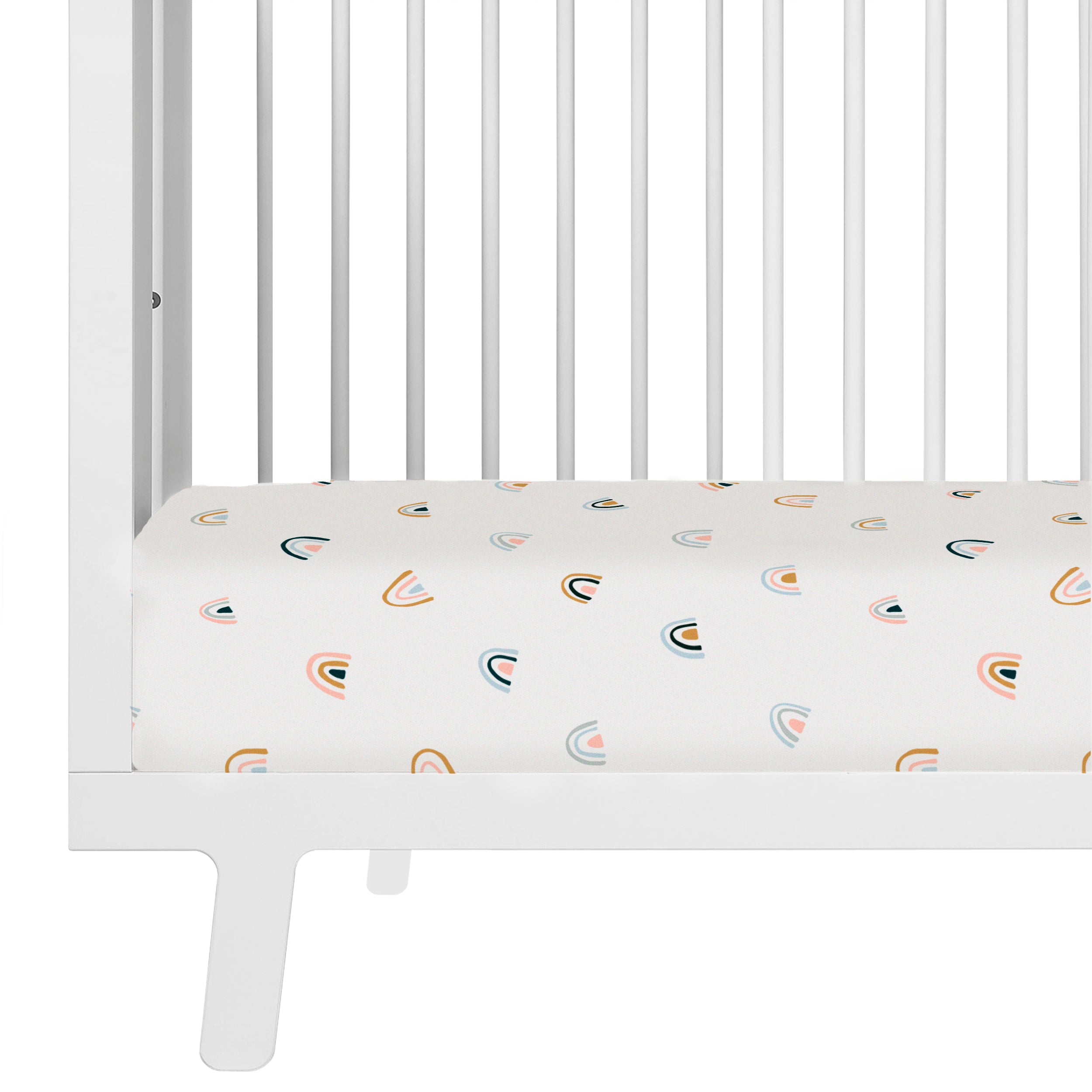 A close-up view of a white baby crib with a mattress covered in a Makemake Organics Crib Fitted Sheet with Pillowcase - Over The Rainbow, set against a white background.