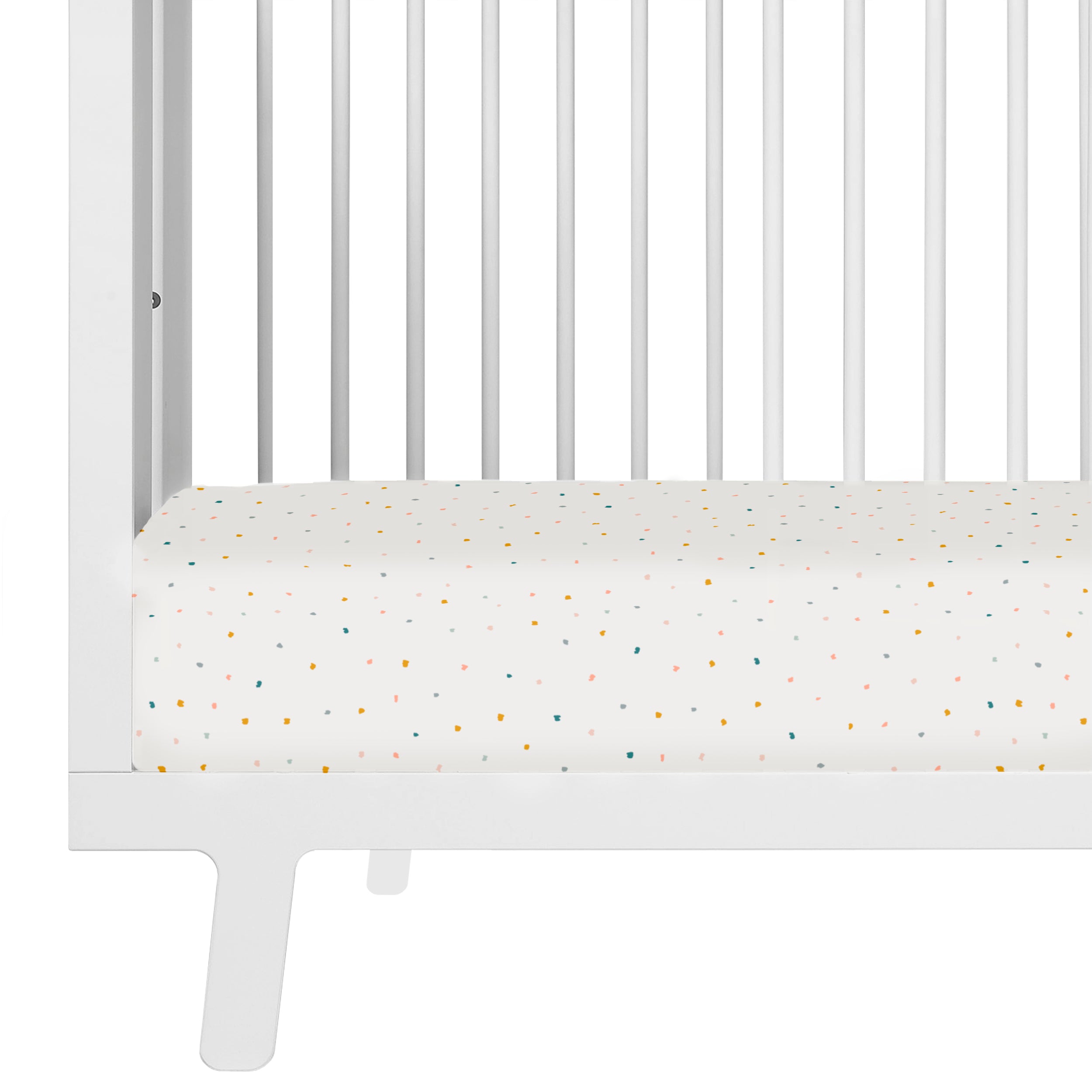 A close-up of a white crib with a mattress covered in a Dotty crib fitted sheet with pillowcase by Makemake Organics, featuring a multicolored pattern. The crib has vertical bars and a simple modern design.