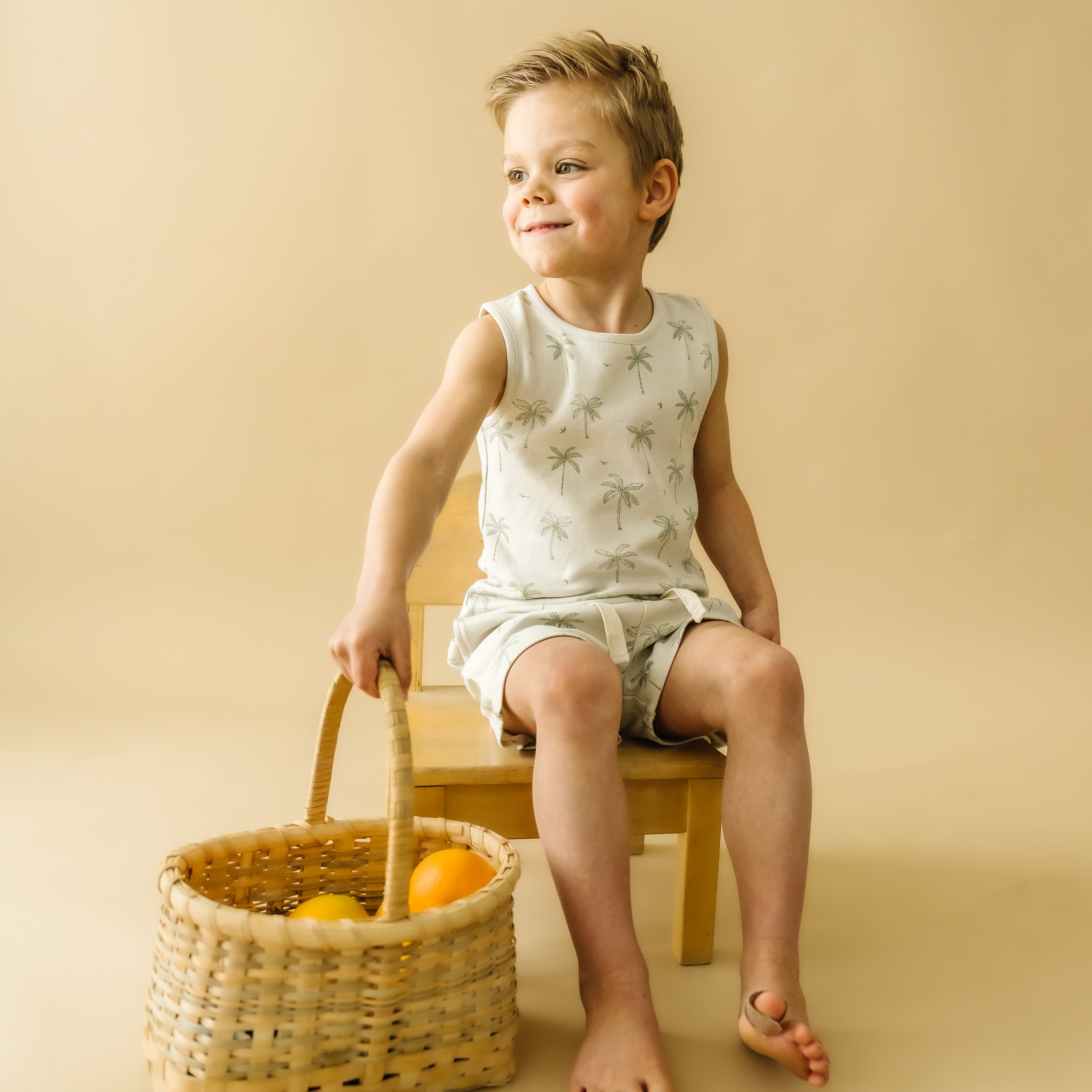 A young boy in a sleeveless shirt and shorts sits on a stool, smiling as he looks to the side, holding a Organic Kids wicker basket filled with oranges. The background is