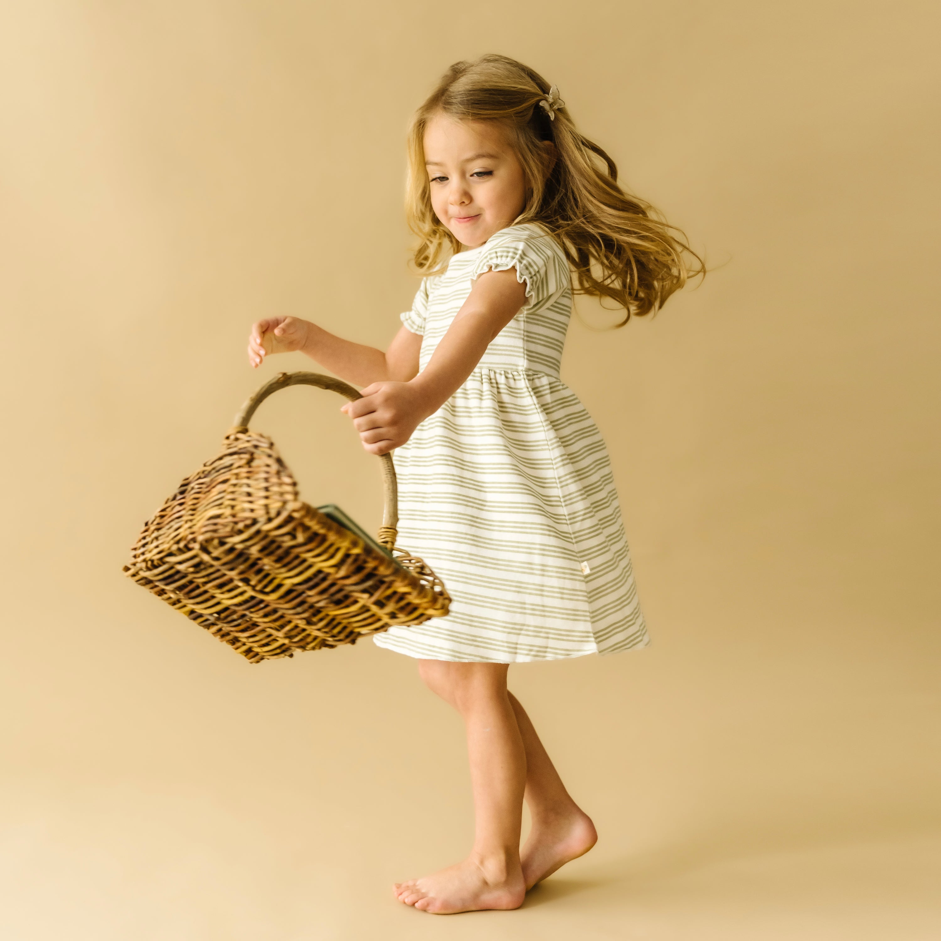 A young girl in an Organic Kids striped dress joyfully leaps while holding a large wicker basket, her bare feet just off the ground and her hair floating gently, against a soft