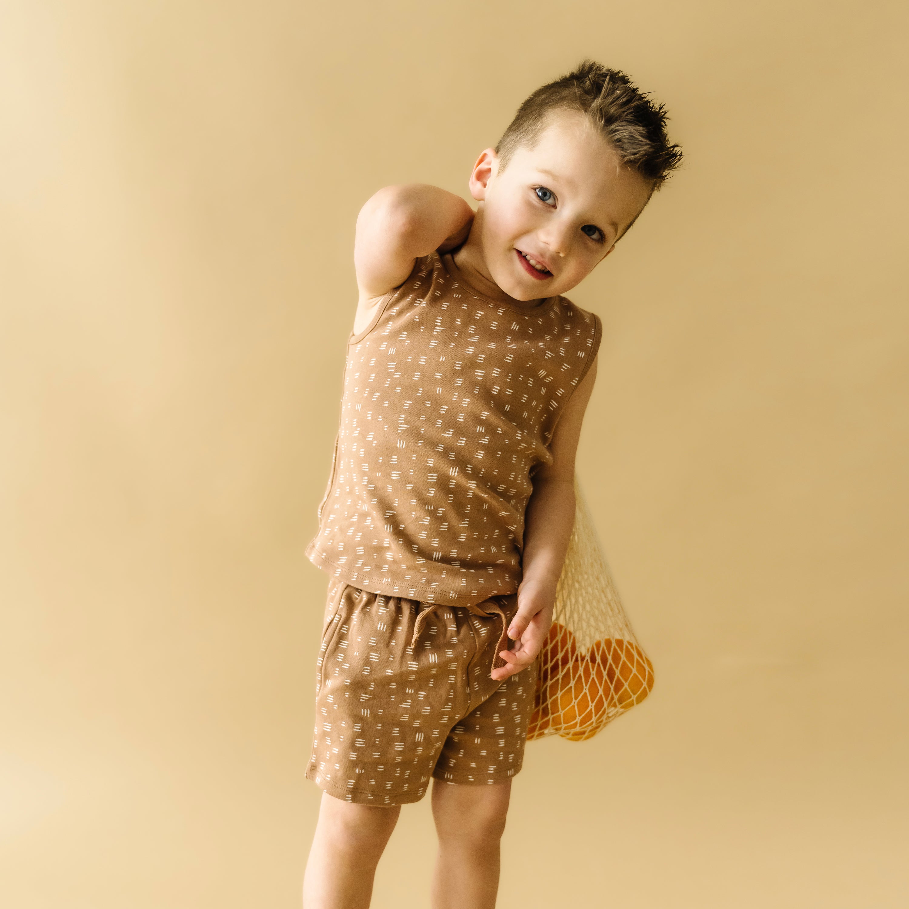 A young boy in a matching brown Organic Tee & Shorts - Strokes set from Organic Kids, playfully posing with one hand on his hip against a beige background. His hair is spiked up.