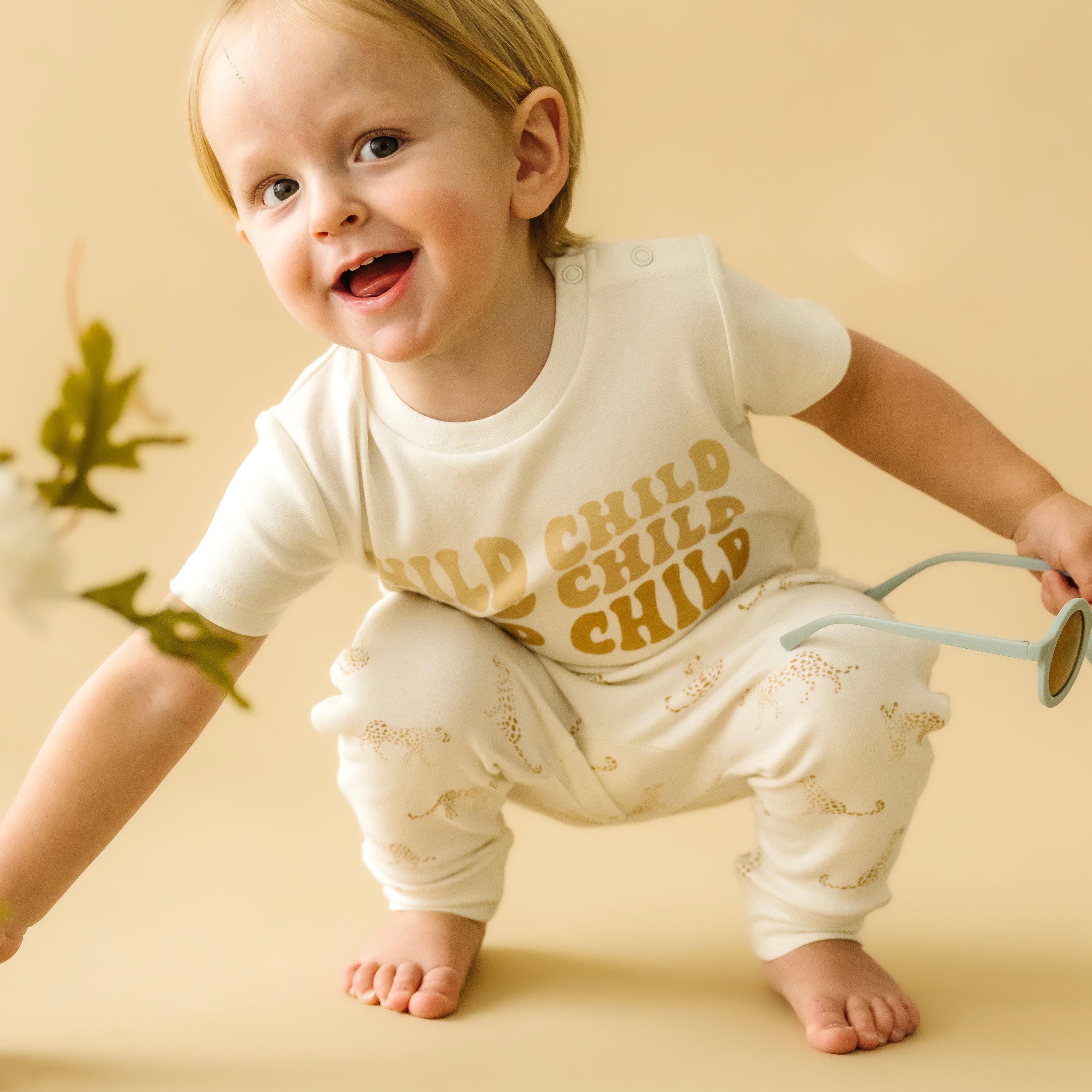 A joyful toddler in a white outfit from Organic Kids with an Organic Tee & Pants Set - Wild Child printed on it, smiling and playing with a blue toy watering can, against a soft beige background.