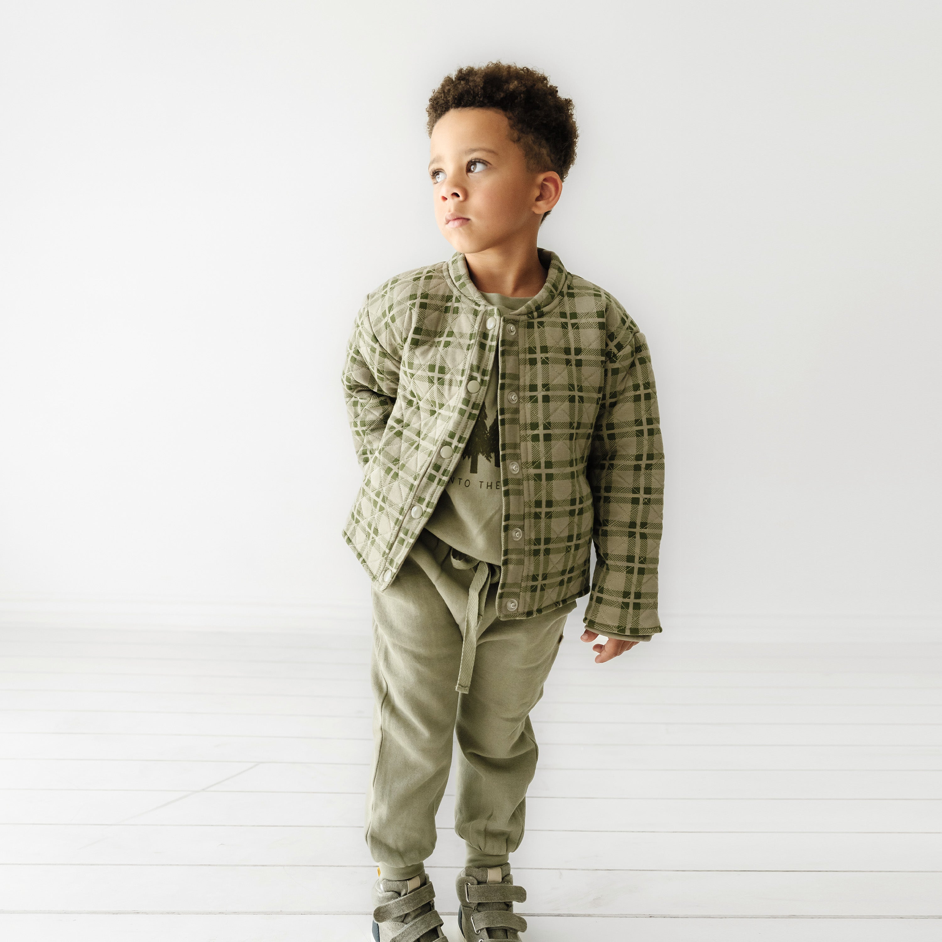 A young boy dressed in a stylish green and brown plaid Organic Merino Wool Buttoned Jacket from Organic Kids, coordinating t-shirt and pants, stands confidently in a white studio setting, looking slightly upwards to his right.