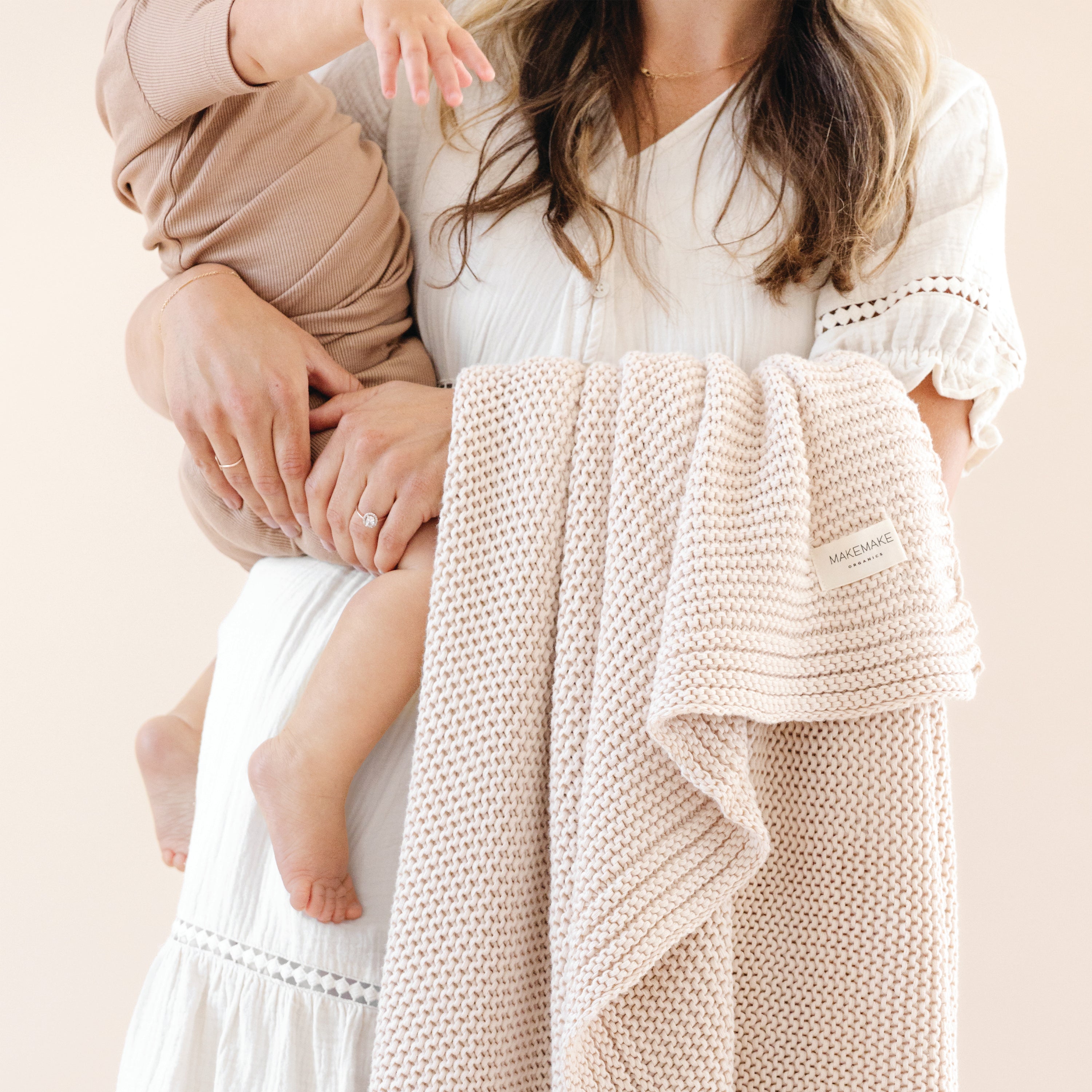 A mother holds her young child, who is wrapped in a Chunky Knit Throw Blanket - Nora Shell by Makemake Organics. Only the lower half of the child's body and the mother's torso are visible, set against a neutral background.