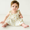 A baby girl with a serious expression sits on the floor wearing a Makemake Organics Organic Smocked Dress in Sunshine, looking directly at the camera.