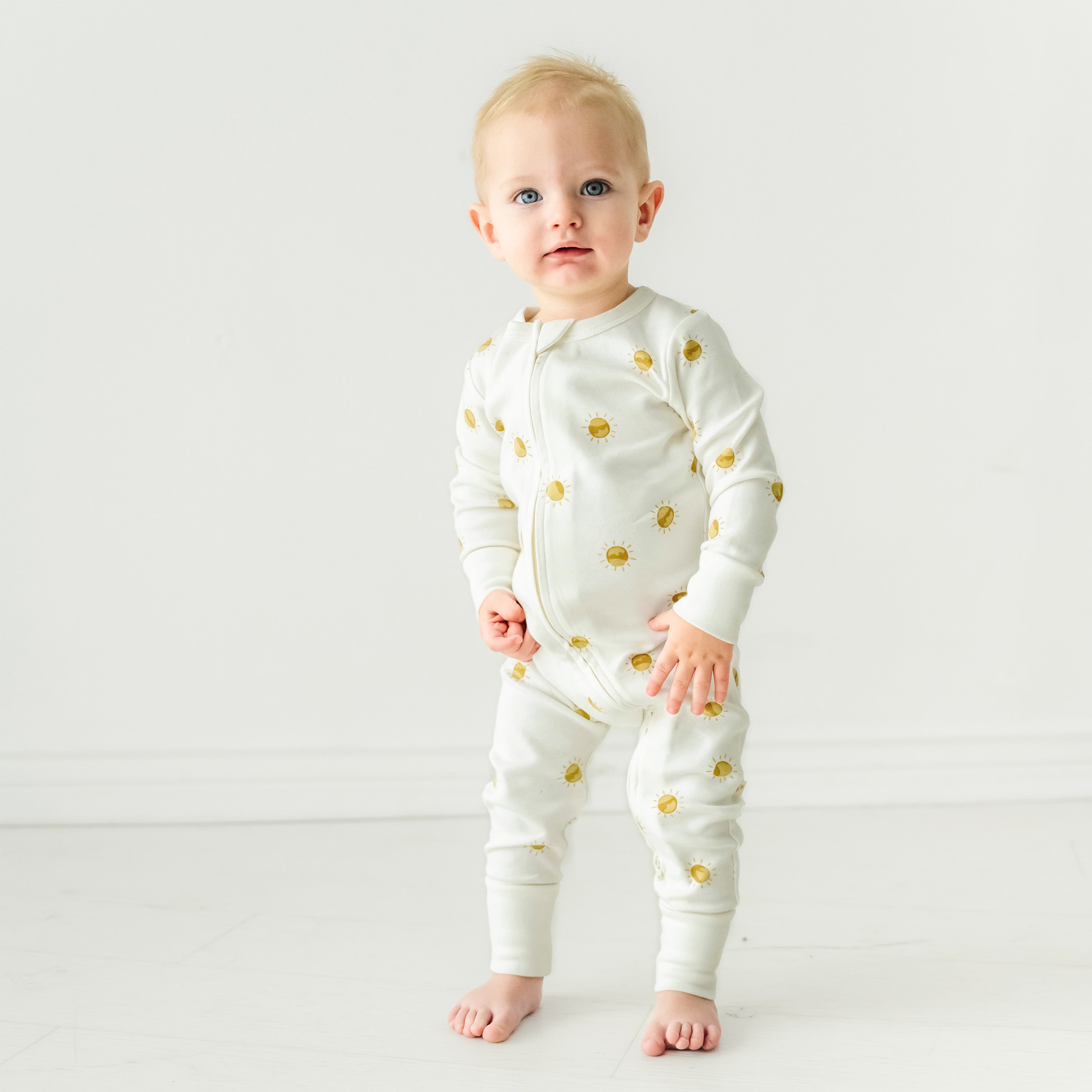 A baby standing on a white background, wearing a white onesie decorated with golden bees, looking directly at the camera with a curious expression. 

becomes

A baby standing on a white background, wearing the Organic 2-Way Zip Romper - Sunshine from Makemake Organics, looking directly at the camera with a curious expression.