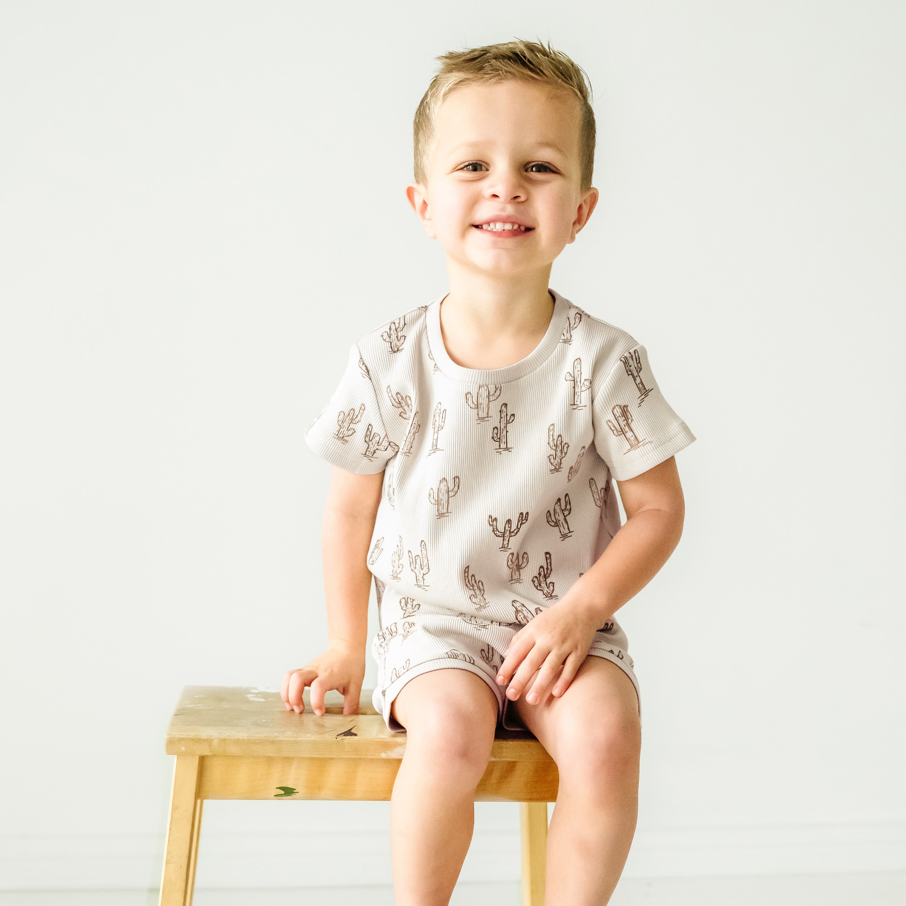 A cheerful young boy sits on a small wooden stool, wearing a white onesie patterned with cacti from the Makemake Organics Organic Tee and Shorties Set. He smiles brightly against a plain white background.