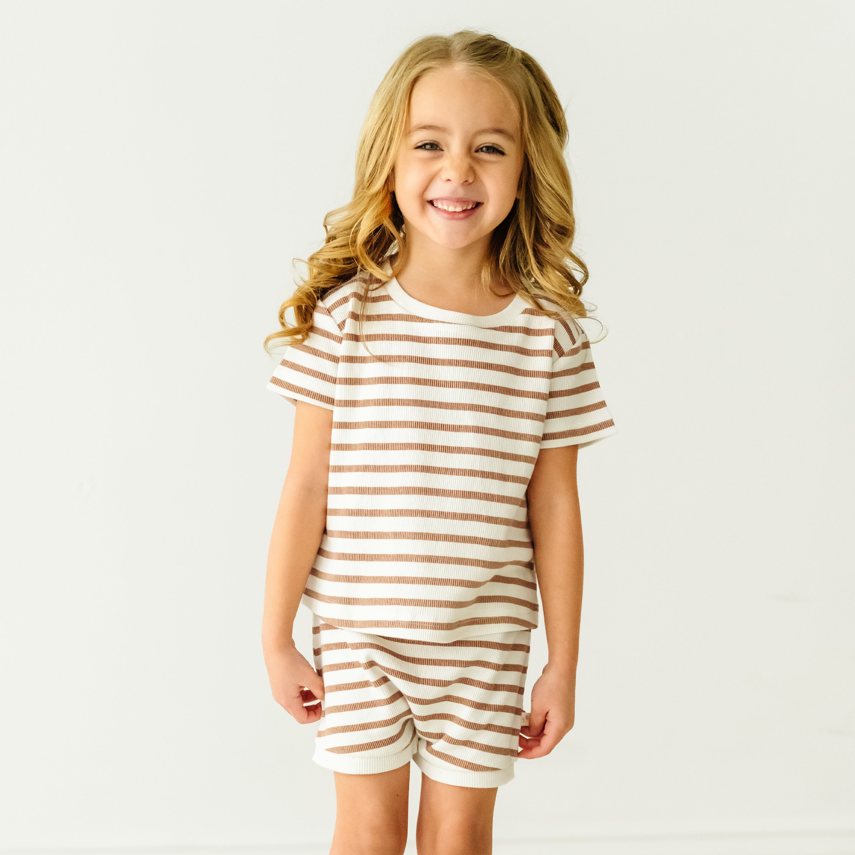 A young girl with curly blonde hair, smiling broadly, wearing the Makemake Organics Organic Tee and Shorties Set in Stripes, standing against a plain white background.