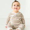 A cheerful toddler with a mohawk hairstyle sits wearing Makemake Organics' Organic Harem Pants in Stripes, smiling joyfully in a bright, white room.