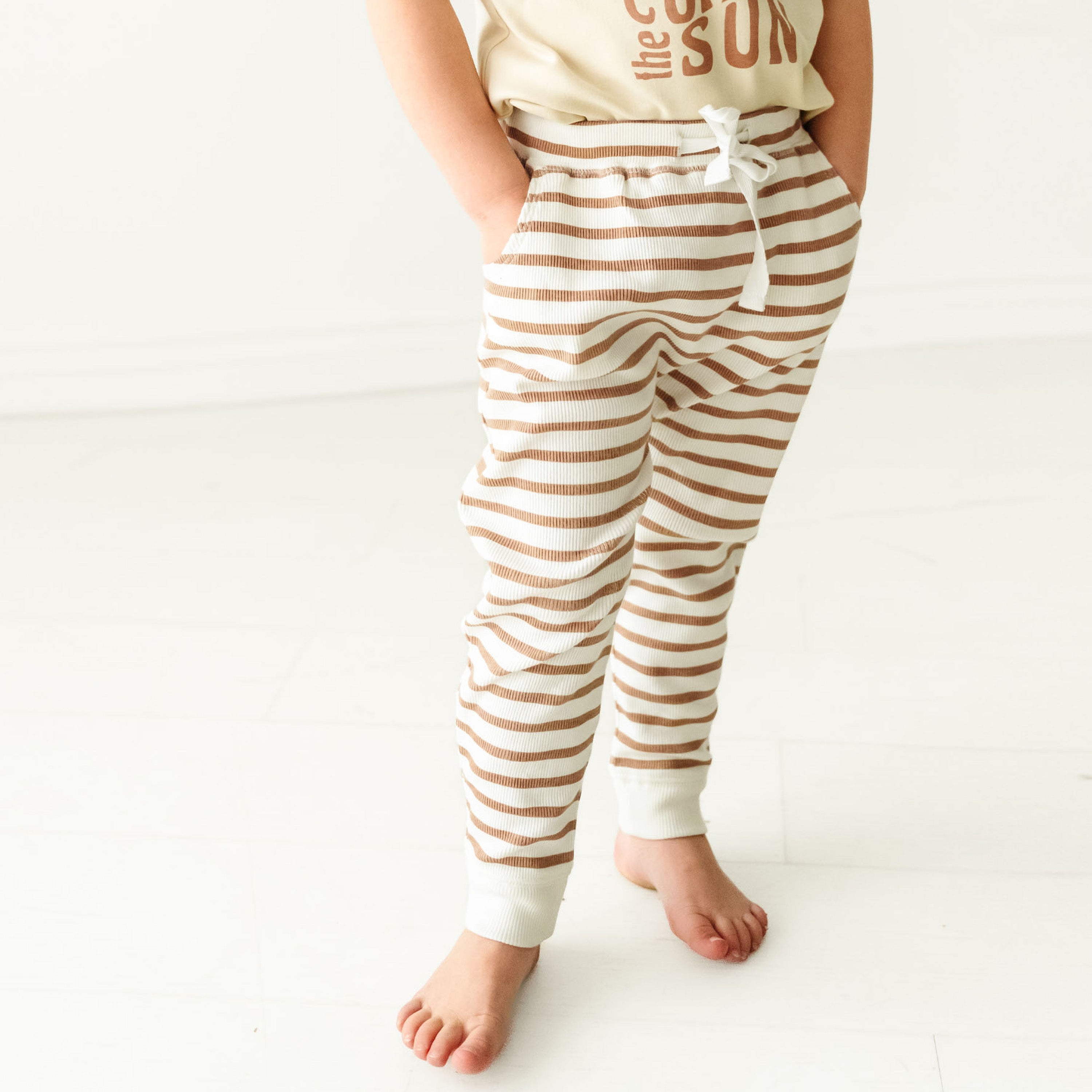 A baby in Makemake Organics' Organic Harem Pants - Stripes stands barefoot on a white floor, with only the lower half of their body visible in the image.