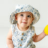A girl toddler with wide eyes wearing an Organic Muslin Bucket Sun Hat in Periwinkle from Makemake Organics and a sleeveless outfit, holding a yellow ball, against a plain white background.