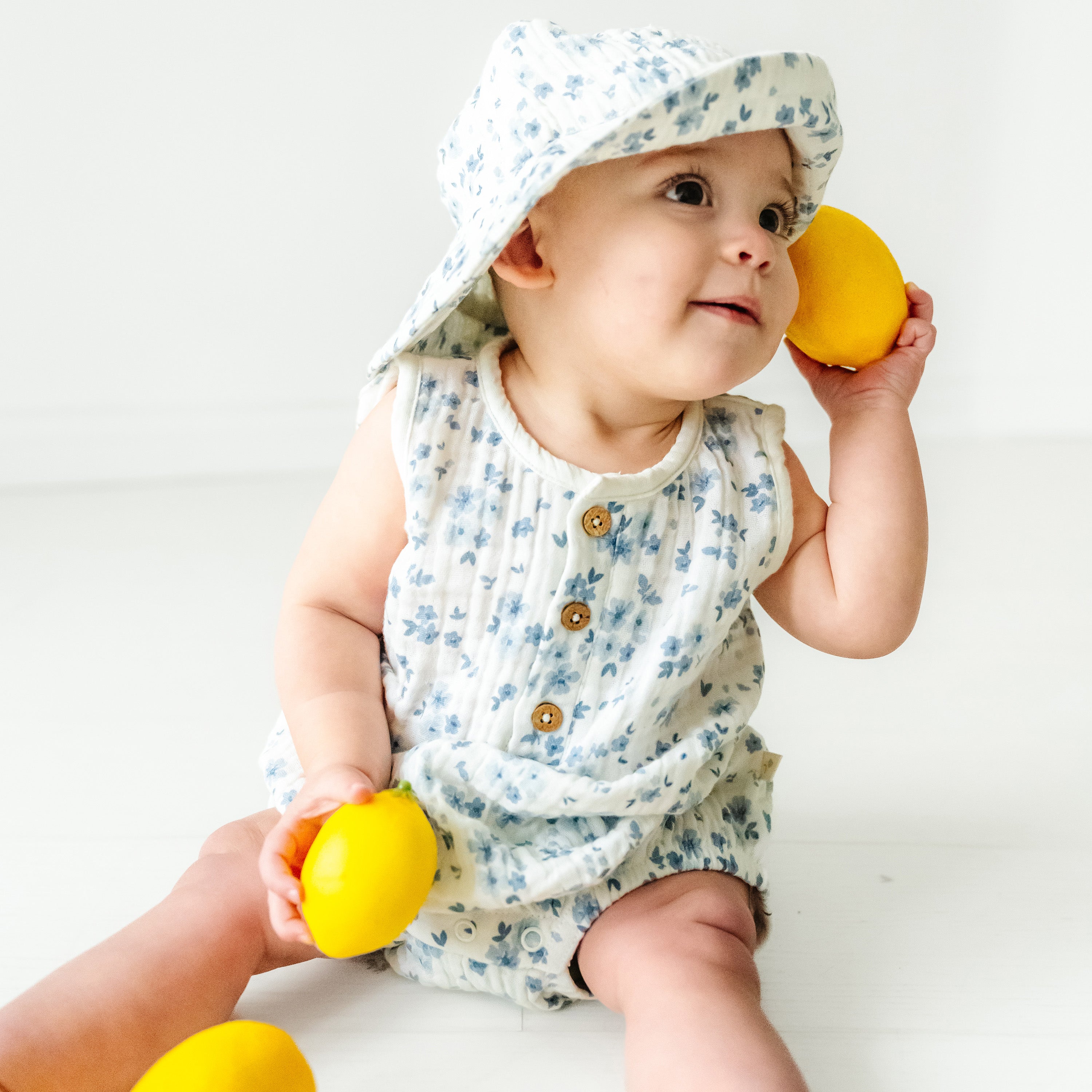 A toddler in a floral outfit and Organic Muslin Bucket Sun Hat - Periwinkle by Makemake Organics sitting and holding yellow balls, looking upwards with a curious expression on a white background.