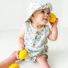 A toddler in a floral outfit and Organic Muslin Bucket Sun Hat - Periwinkle by Makemake Organics sitting and holding yellow balls, looking upwards with a curious expression on a white background.