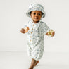 A baby girl wearing the Makemake Organics Organic Muslin Short Bubble Romper in Periwinkle and matching bucket hat walking joyfully while holding a small yellow toy, set against a plain white background.