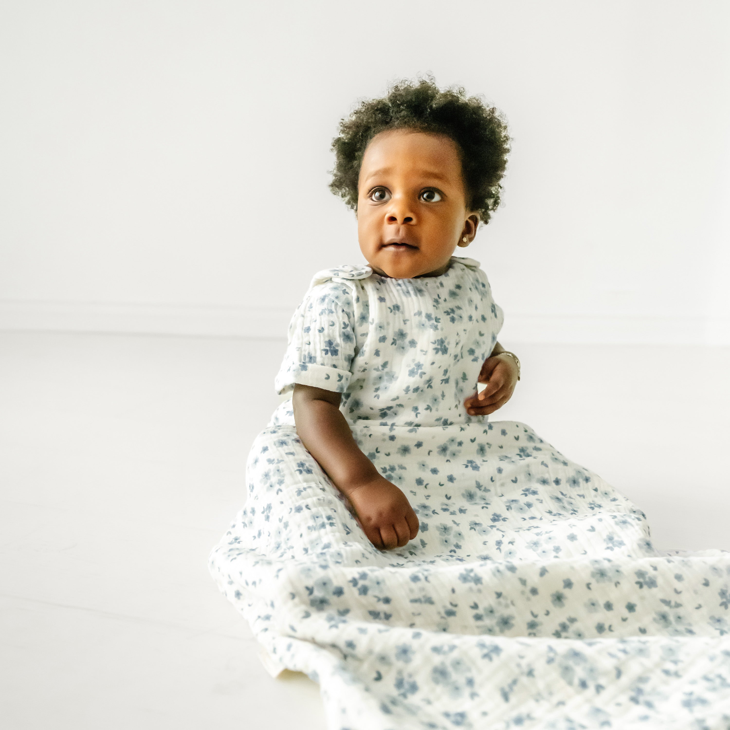 A toddler with curly hair sits on a Makemake Organics Muslin Wearable Blanket in Periwinkle, wearing a floral dress, looking up with a curious expression in a bright, airy room.