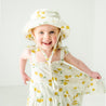 A joyful baby girl wearing a Makemake Organics Organic Linen Bucket Sun Hat in Citron and matching sun hat, smiling brightly in a well-lit room with a clean, white backdrop.