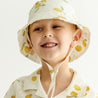 A joyful toddler wearing a Makemake Organics Organic Linen Bucket Sun Hat - Citron and matching shirt, smiling broadly against a light background.
