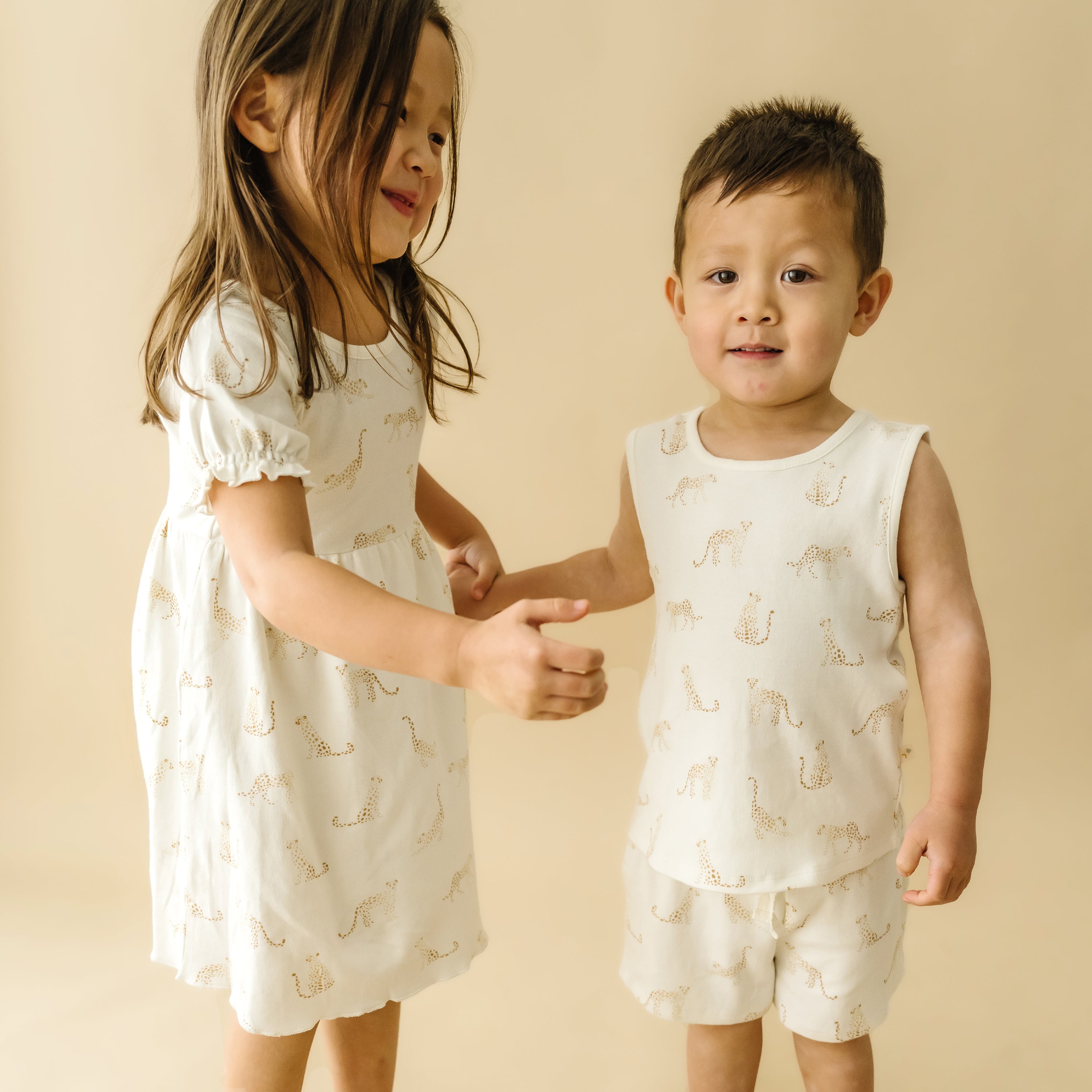 Two young children, a girl in a white dress and a boy in a matching white outfit with giraffe prints from Organic Kids, are holding hands and smiling in front of a beige background.