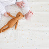 A baby lying on a Makemake Organics multicolored polka dot blanket, holding a brown stuffed bunny toy next to their bare feet.