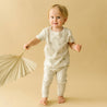 A toddler with light hair, dressed in a white and beige Organic Kids outfit with a palm leaf pattern, stands smiling and holding a paper fan in a beige studio background.