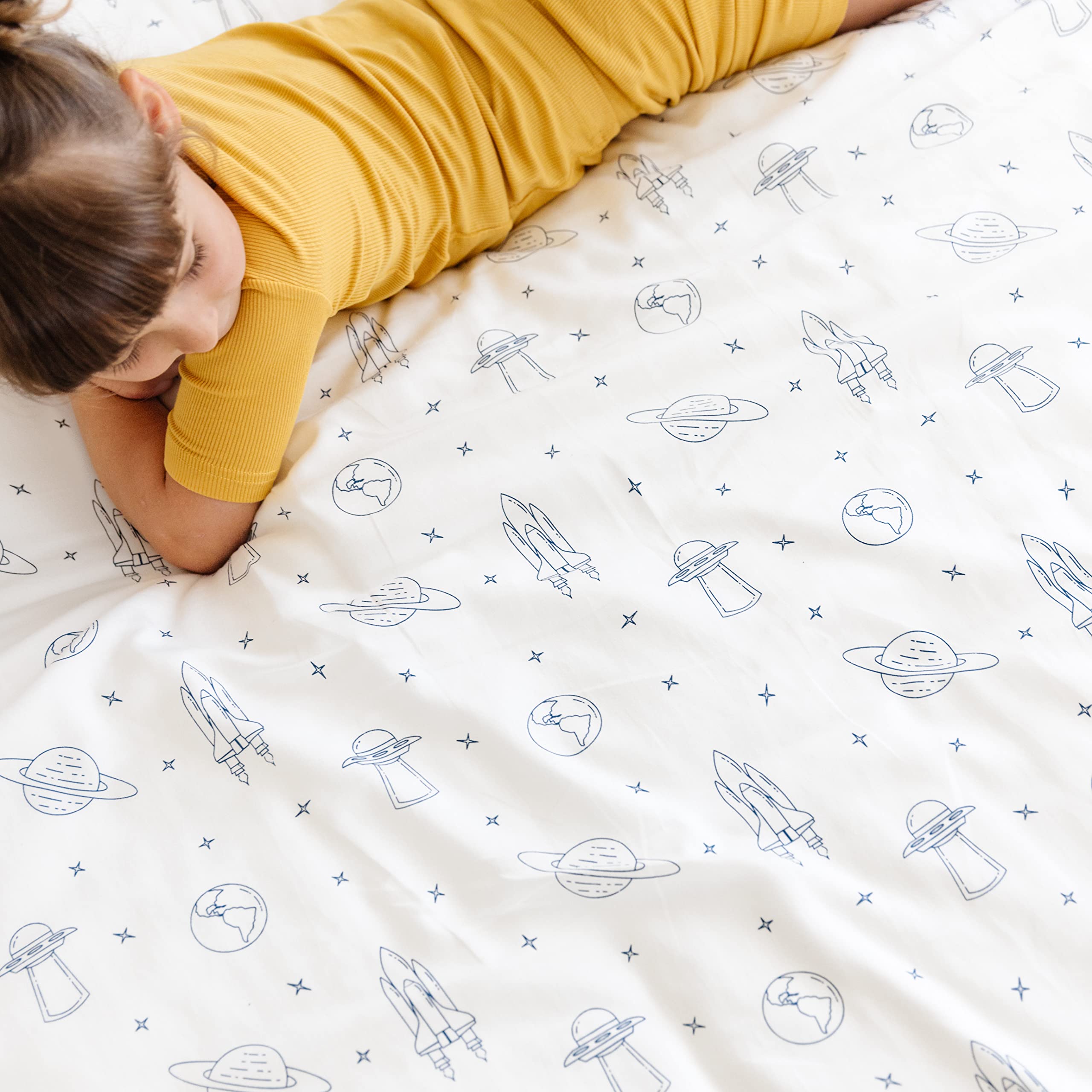 A child in a yellow top is lying face down on a bed, resting their head on their arms, with a Makemake Organics Celestial Crib Fitted Sheet with Pillowcase featuring a space-themed pattern with rockets and planets.